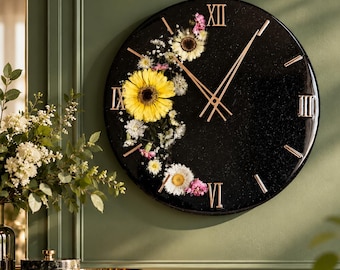 May include: A round black wall clock with a glossy finish and embedded pressed flowers in yellow and white. The clock face features gold-colored Roman numerals at the three, six, nine, and twelve o'clock positions, with thin gold hands for timekeeping.