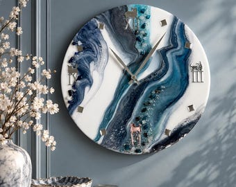 May include: A round wall clock featuring a white, navy blue, and teal resin pour design with a geode-inspired center. The clock face includes silver-toned hands, square hour markers, and three metallic animal silhouettes at the XII, IX, and III positions.