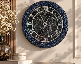 May include: A round wall clock featuring a black and white marbled face with silver Roman numerals and hands. The clock is framed by a textured border of deep blue crushed glass or stone fragments, creating a high-contrast circular design.