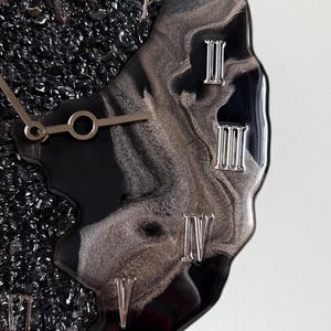 May include: A wall clock featuring a black and silver marbled resin surface with embedded black crushed glass. The clock face displays silver Roman numerals and metallic hands, showcasing a fluid, irregular edge design with a glossy finish.