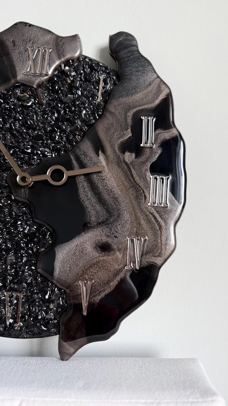 May include: A wall clock featuring a black and silver marbled resin surface with embedded black crushed glass. The clock face displays silver Roman numerals and metallic hands, showcasing a fluid, irregular edge design with a glossy finish.