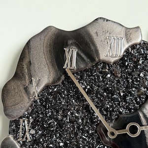 May include: A wall clock featuring a center filled with small, dark, reflective black stones. The outer edge has irregular, smooth, metallic silver sections with raised Roman numerals in silver. Two thin, silver clock hands extend from the center.