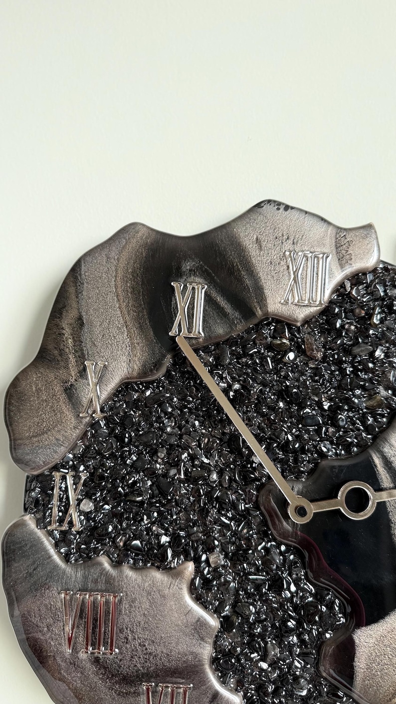 May include: A wall clock featuring a center filled with small, dark, reflective black stones. The outer edge has irregular, smooth, metallic silver sections with raised Roman numerals in silver. Two thin, silver clock hands extend from the center.