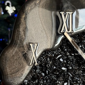 May include: A wall clock featuring a glossy, marbled black and silver resin border with metallic silver Roman numerals IX, X, and XI. The center of the clock is filled with small, dark, reflective crushed stone fragments and a silver clock hand.