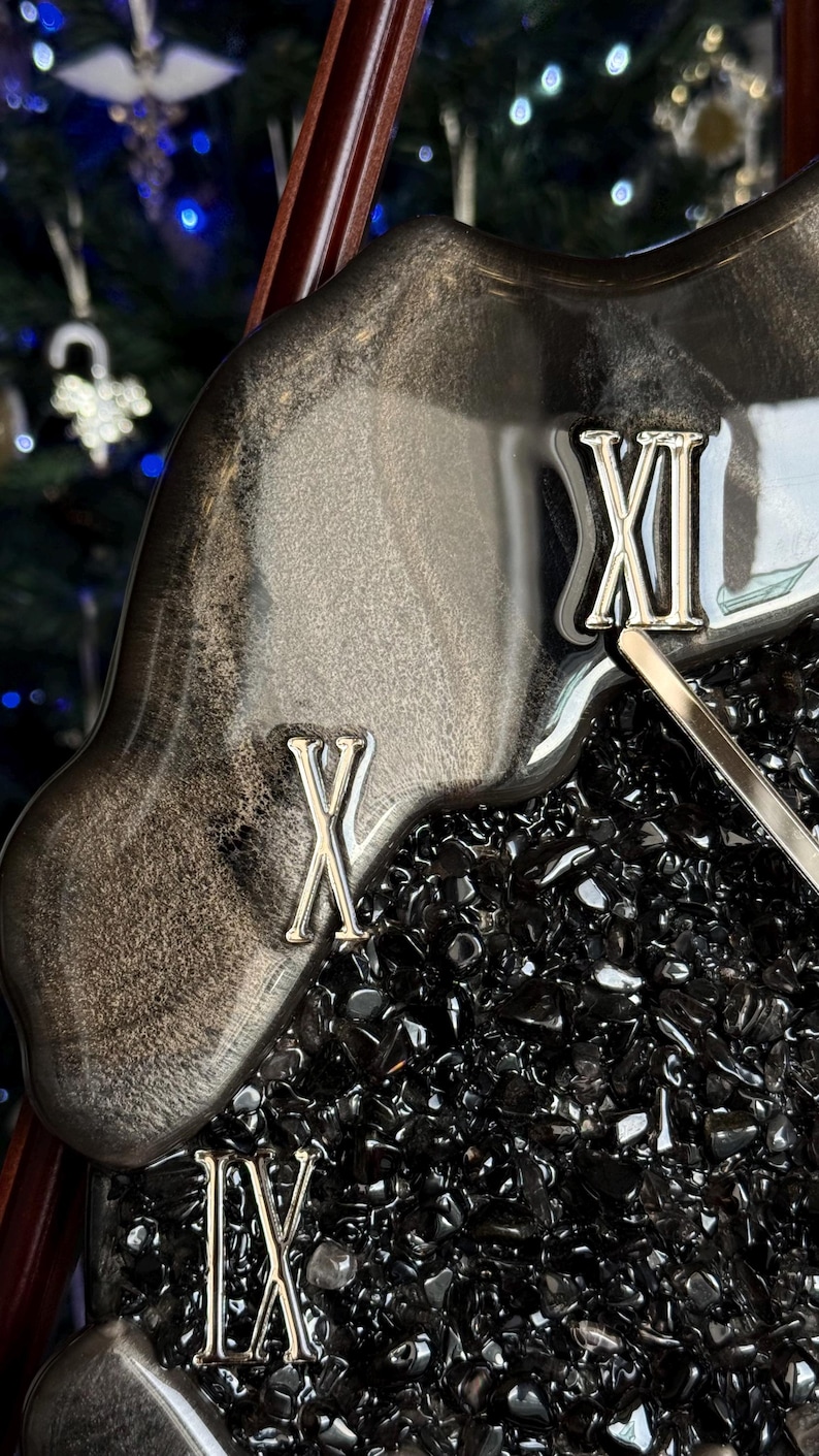 May include: A wall clock featuring a glossy, marbled black and silver resin border with metallic silver Roman numerals IX, X, and XI. The center of the clock is filled with small, dark, reflective crushed stone fragments and a silver clock hand.