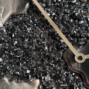 May include: A wall clock featuring a textured center filled with small, polished black stones. The clock face has a smooth, dark gray border with raised silver Roman numerals. Two thin, straight silver clock hands extend from the center.