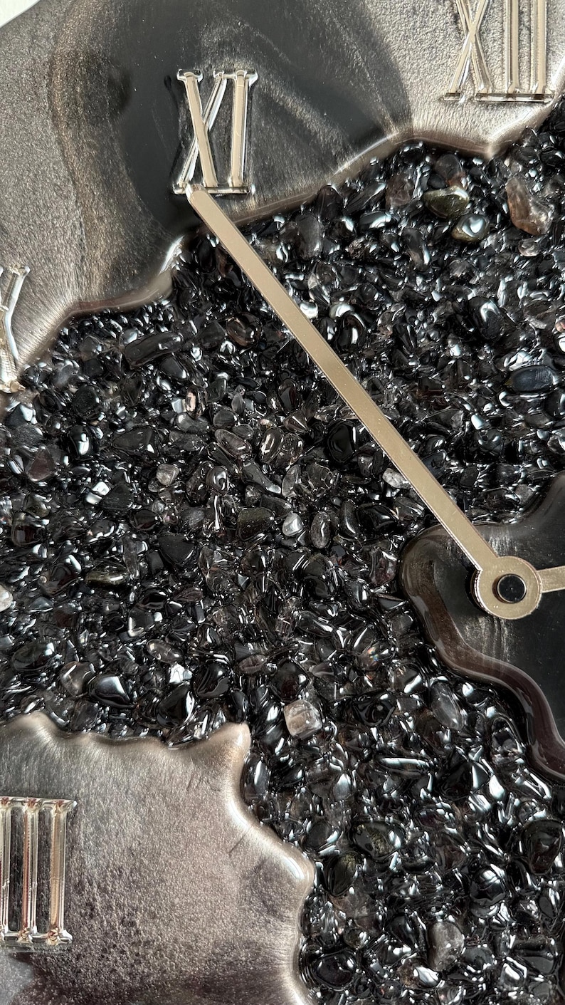 May include: A wall clock featuring a textured center filled with small, polished black stones. The clock face has a smooth, dark gray border with raised silver Roman numerals. Two thin, straight silver clock hands extend from the center.