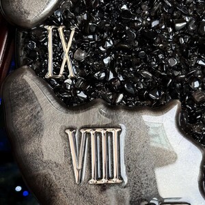 May include: A wall clock face featuring a glossy, marbled black and silver resin border with raised metallic Roman numerals IX, VIII, and VII. The center is filled with a dense layer of small, polished black obsidian-like stone chips.