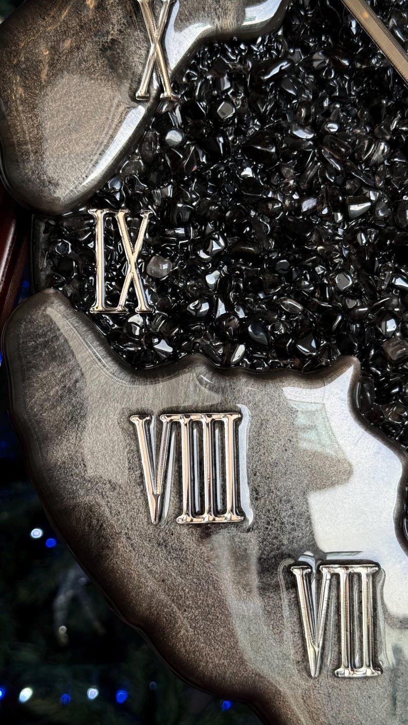 May include: A wall clock face featuring a glossy, marbled black and silver resin border with raised metallic Roman numerals IX, VIII, and VII. The center is filled with a dense layer of small, polished black obsidian-like stone chips.