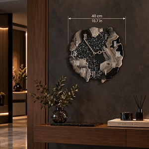 May include: A circular wall clock with an irregular, organic edge measuring 15.7 inches in diameter. The clock face features a central section of black textured material, surrounded by swirling gray and black resin, with gold Roman numerals and hands.