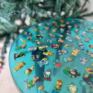 May include: A turquoise resin table top with a variety of colorful figurines embedded in the surface. The figurines are mostly cartoon characters and animals.