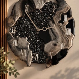 May include: A wall clock with an irregular, organic shape featuring a black and silver marbled resin finish. The center is filled with black crushed glass, and the face displays silver Roman numerals from I to XII with thin silver clock hands.