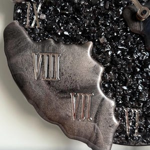 May include: A wall clock featuring a textured black center filled with small, dark, reflective stone-like fragments. The outer edge is composed of a smooth, metallic silver resin with raised Roman numerals VII, VIII, IX, and X in silver.