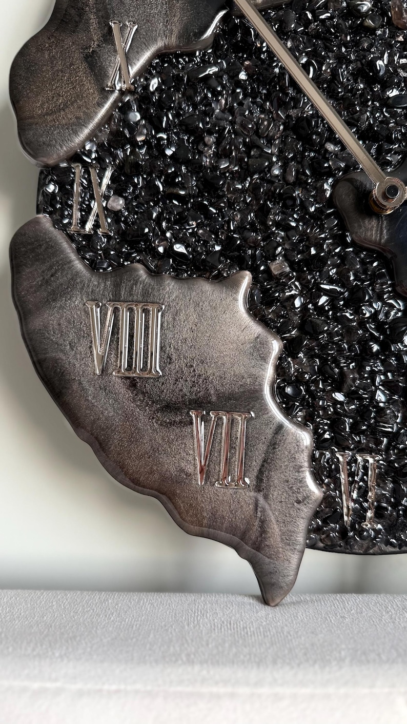 May include: A wall clock featuring a textured black center filled with small, dark, reflective stone-like fragments. The outer edge is composed of a smooth, metallic silver resin with raised Roman numerals VII, VIII, IX, and X in silver.