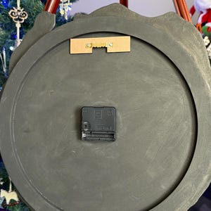 May include: The back of a circular wall clock featuring a dark gray, matte-finished frame. A black plastic battery-operated clock mechanism is centered on the back, and a rectangular wooden mounting bracket with a gold-toned metal sawtooth hanger is attached near the top edge.