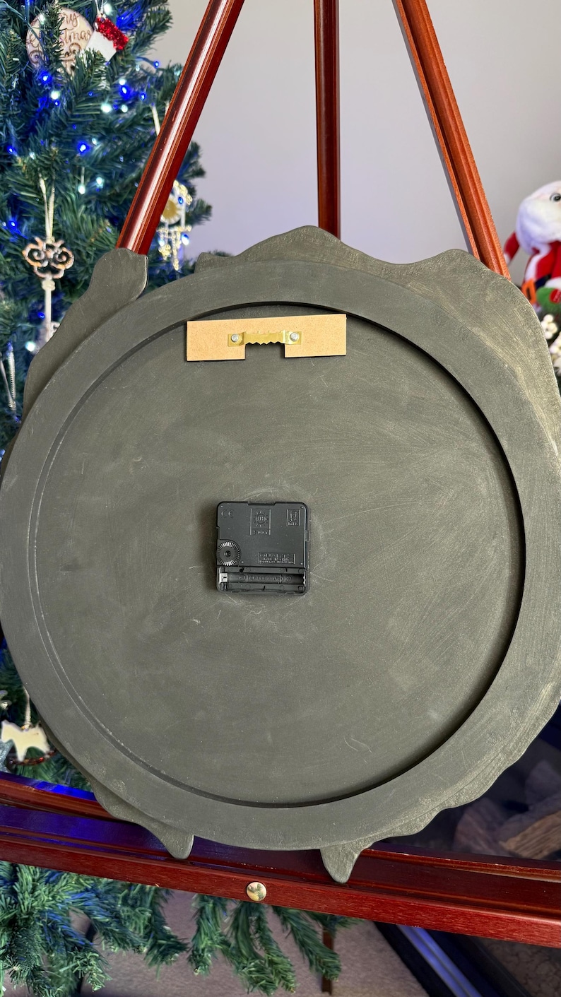 May include: The back of a circular wall clock featuring a dark gray, matte-finished frame. A black plastic battery-operated clock mechanism is centered on the back, and a rectangular wooden mounting bracket with a gold-toned metal sawtooth hanger is attached near the top edge.