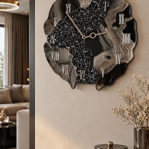 May include: A wall clock with an irregular, organic shape featuring a black and silver marbled resin finish. The center contains a textured black crystalline inlay. It displays silver Roman numerals and thin, gold-toned metal clock hands.
