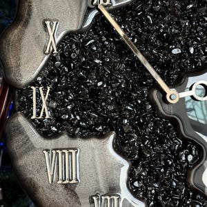 May include: A wall clock featuring a black resin frame with a swirling metallic silver finish. The center is filled with small, polished black stones. Silver Roman numerals are embedded along the edge, and thin silver hands indicate the time.
