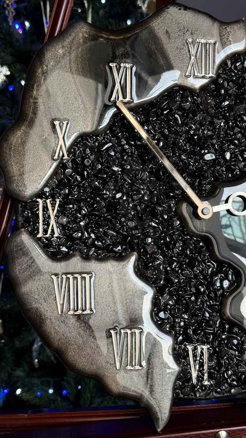 May include: A wall clock featuring a black resin frame with a swirling metallic silver finish. The center is filled with small, polished black stones. Silver Roman numerals are embedded along the edge, and thin silver hands indicate the time.
