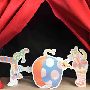 May include: A whimsical puppet show scene with a red curtain backdrop. The stage features three colorful cutouts: a clown doing a handstand, a large pumpkin-shaped prop, and a cat-clown character. The scene evokes a playful, circus-like atmosphere.