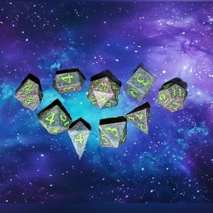 Puede incluir: Un conjunto de nueve dados poliédricos para juegos de mesa. Los dados son de color gris oscuro con purpurina, números verde neón y detalles en rosa. El fondo es una galaxia púrpura y azul.