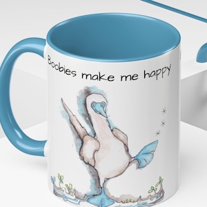 May include: White ceramic mug with a blue interior and handle. The mug features a cartoon illustration of a blue-footed booby and the text "Boobies make me happy" in black.