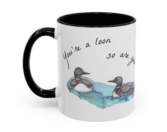 May include: White ceramic mug with a black handle and interior. The mug features a watercolor illustration of two loons on a blue water background, with the handwritten text "You're a loon so are you".