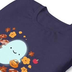Pu&ograve; includere: Una maglietta blu navy con un fantasma bianco che tiene un cestino di zucca. Il fantasma &egrave; circondato da foglie autunnali arancioni e marroni e funghi.