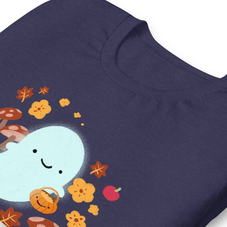 Pu&ograve; includere: Una maglietta blu navy con un fantasma bianco che tiene un cestino di zucca. Il fantasma &egrave; circondato da foglie autunnali arancioni e marroni e funghi.