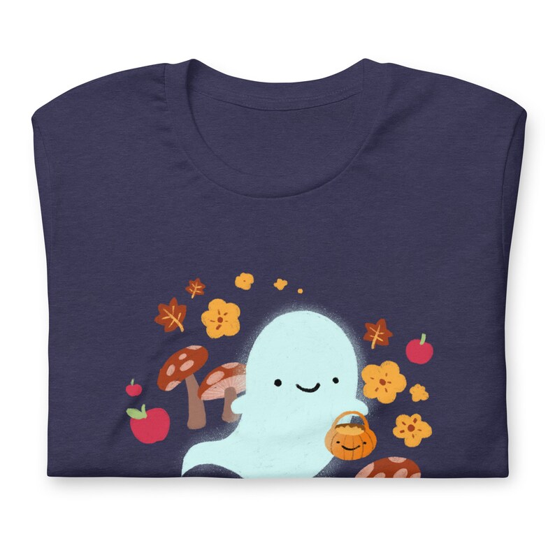 Pu&ograve; includere: Una maglietta blu navy con un simpatico fantasma che tiene un secchiello di zucca. Il fantasma &egrave; circondato da foglie autunnali, funghi e fiori.