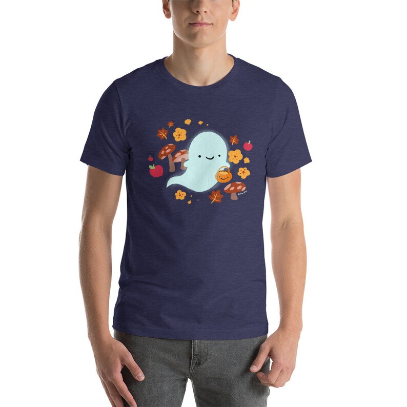 Pu&ograve; includere: Una maglietta blu navy con un simpatico fantasma dei cartoni animati che tiene un secchiello di zucca. Il fantasma &egrave; circondato da foglie autunnali, funghi e mele.