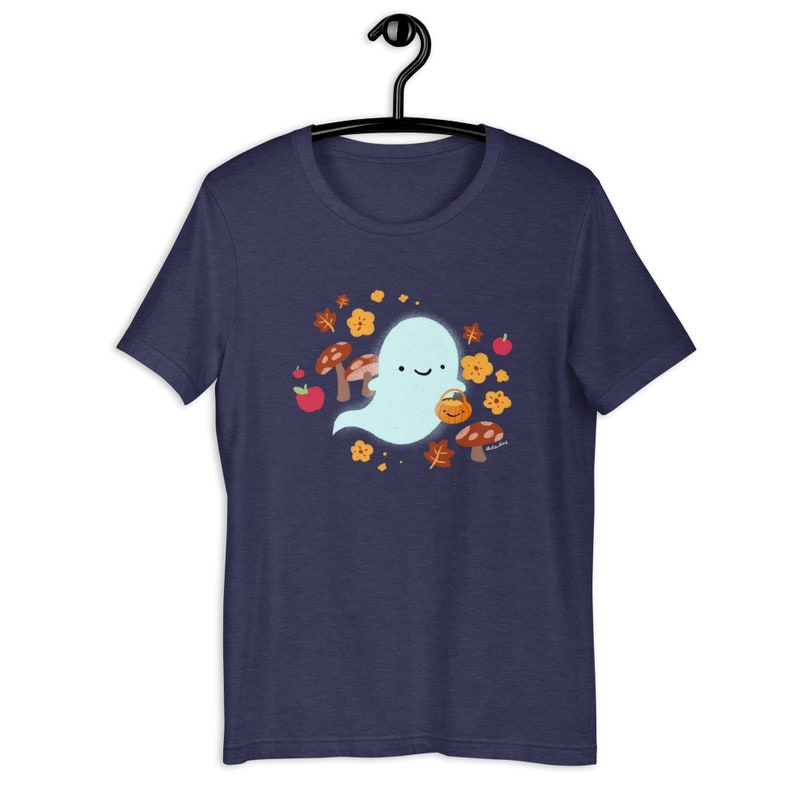 Pu&ograve; includere: Una maglietta blu navy con un simpatico fantasma dei cartoni animati che tiene un secchiello di zucca. Il fantasma &egrave; circondato da foglie autunnali, funghi e mele.