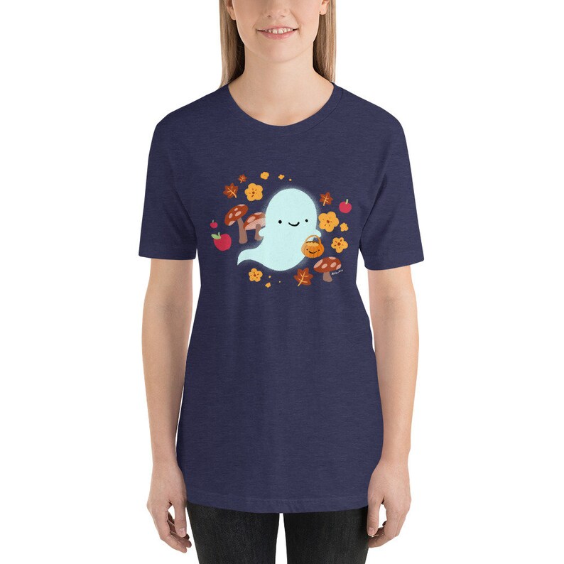 Pu&ograve; includere: Una maglietta blu navy con un simpatico fantasma dei cartoni animati che tiene un secchiello di zucca. Il fantasma &egrave; circondato da foglie autunnali, funghi e mele.
