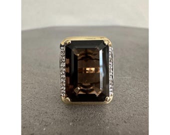 Puede incluir: Anillo con una gran piedra de cuarzo ahumado de corte rectangular. El anillo tiene una banda dorada con un engaste rectangular y un borde de pequeñas piedras brillantes. El cuarzo es de color marrón oscuro.
