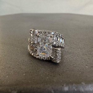 Puede incluir: Un anillo plateado con una gran gema rectangular y transparente como punto focal. El anillo está incrustado con gemas claras más pequeñas, creando un efecto brillante. El diseño presenta un estilo envolvente con múltiples bandas.