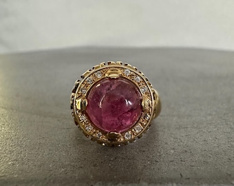 Può includere: Un anello d'oro con una grande gemma rosa ovale al centro. La gemma è circondata da un alone di piccole gemme trasparenti. L'anello presenta intricati dettagli dorati attorno alla gemma e al gambo.
