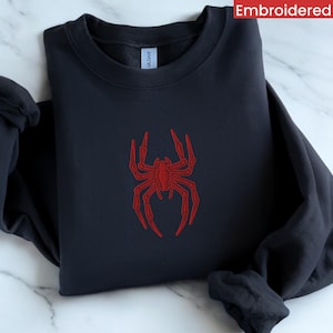 May include: A dark blue sweatshirt featuring a red embroidered spider design. The spider has eight legs and a body with a red outline. The word "Embroidered" is printed in white in the top right corner of the image.