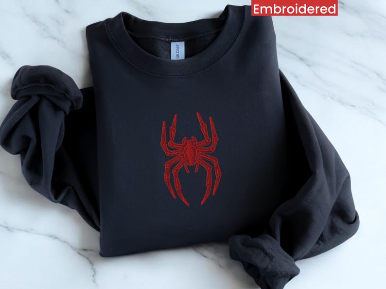 May include: A dark blue sweatshirt featuring a red embroidered spider design. The spider has eight legs and a body with a red outline. The word "Embroidered" is printed in white in the top right corner of the image.
