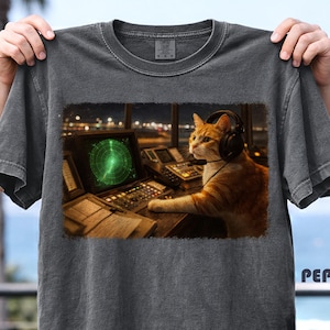 May include: Dark gray t-shirt featuring a graphic of an orange tabby cat wearing headphones and operating a radar system. The cat is in a control room with a green radar screen. The word "PEPPER" is printed on the bottom right.