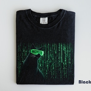 May include: Black t-shirt featuring a cat wearing glowing green glasses, set against a green digital rain design. The shirt is folded and has a tag at the collar. The word "Black" is printed on the bottom right.