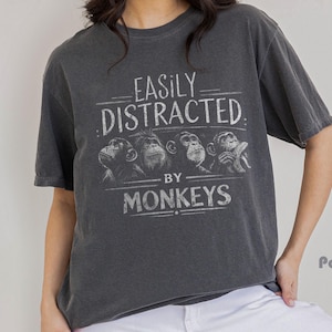 May include: Dark gray t-shirt with the text "EASILY DISTRACTED BY MONKEYS" in white. The shirt features a graphic of four monkey illustrations. The shirt is a casual, unisex style.