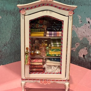 May include: A miniature white wooden cabinet with a glass door, decorated with small pink roses and a colorful braided trim. The interior shelves are filled with stacks of folded miniature textiles, fabrics, and several small glass perfume bottles.