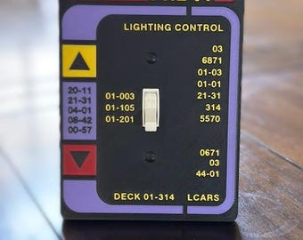 Könnte beinhalten: Eine schwarze und lila Lichtschalterabdeckung mit dem Text "PNL 01" und "LIGHTING CONTROL". Die Abdeckung hat einen Kippschalter und verschiedene Zahlen und Beschriftungen, darunter "DECK 01-314" und "LCARS".