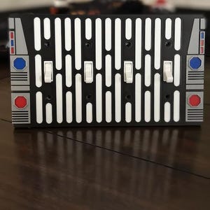 May include: A black light switch plate with a white and black Star Wars inspired design. The plate has six toggle switches and red and blue accents.