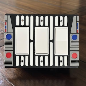 May include: A black light switch cover with three white switches. The cover has a geometric design with red, blue, and gray accents. The design is reminiscent of a science fiction theme.