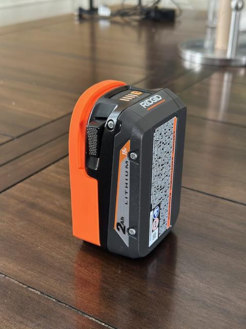 May include: An orange and black Ridgid 18V 2Ah Lithium battery. The battery has a black label with the text "RIDGID" and "2Ah Lithium" printed on it.
