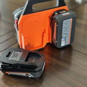 Pode incluir: Um carregador de bateria preto e laranja com duas ranhuras para baterias. O carregador tem uma al&ccedil;a e o texto "RIDGID" no lado. Uma bateria preta est&aacute; localizada ao lado do carregador.