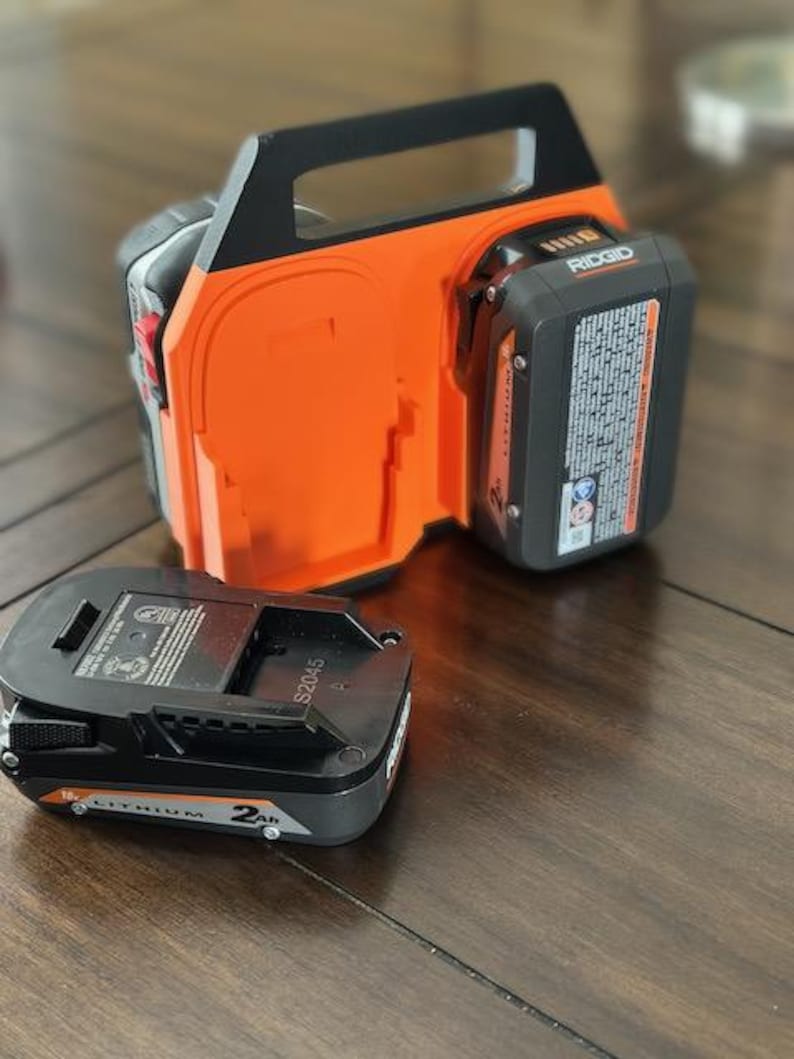 Pode incluir: Um carregador de bateria preto e laranja com duas ranhuras para baterias. O carregador tem uma al&ccedil;a e o texto "RIDGID" no lado. Uma bateria preta est&aacute; localizada ao lado do carregador.