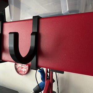 May include: A black U-shaped hook attached to a red metal beam, designed for hanging items. The beam is part of a storage system, with clear plastic bins visible above. A heat gun and other tools are also visible.