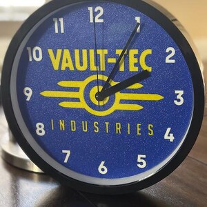 May include: A blue and yellow Vault-Tec Industries clock with black hands and white numbers. The clock has a black frame and a glittery blue face.